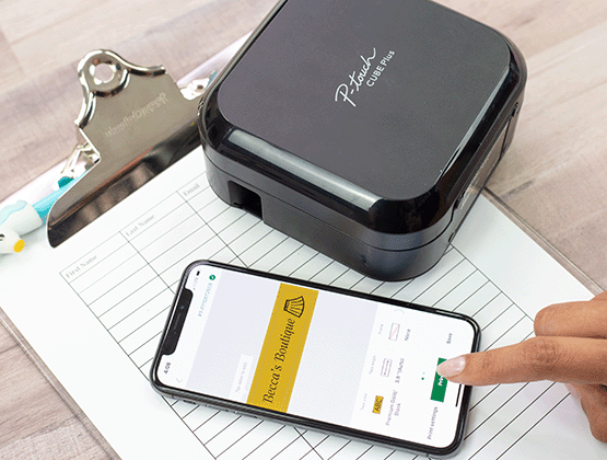 Person creating a label with "Becca's Boutique" on Design&Print app. P-touch CUBE Plus on a clipboard. 