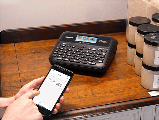 Person printing a barcode label with iPrint&Label app on a P-touch machine sitting on a counter with candles to the right. 