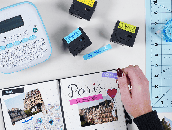  A label maker with yellow and blue tapes next to a journal page labeled "Paris" with cut-out images being added.